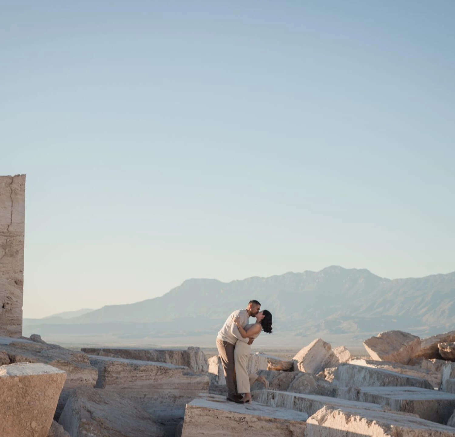 Pareja besándose entre formaciones rocosas al atardecer, con montañas al fondo, celebrando su amor en un paisaje natural y único.