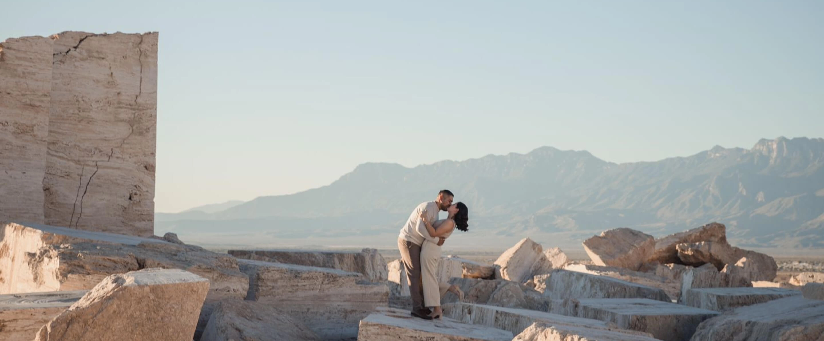 Pareja besándose entre formaciones rocosas al atardecer, con montañas al fondo, celebrando su amor en un paisaje natural y único.