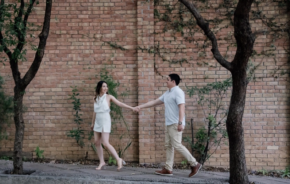 Pareja caminando de la mano frente a una pared de ladrillo, rodeados de árboles, disfrutando un paseo juntos.