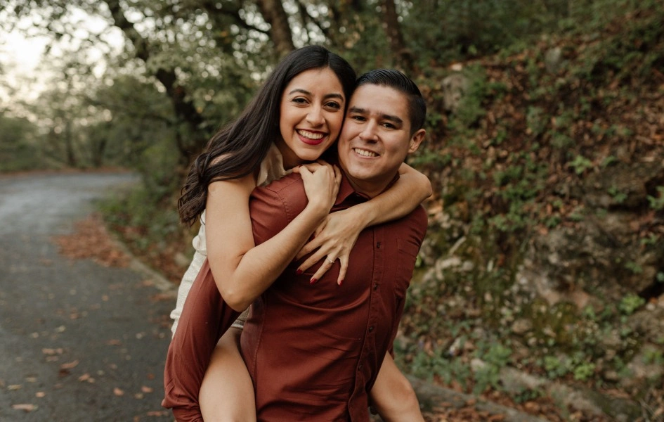 Pareja sonriendo mientras ella lo abraza por la espalda en un camino rodeado de naturaleza, transmitiendo felicidad y complicidad.